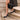 A person wearing black pointed-toe high heels and carrying a black shoulder bag with a gold clasp, standing on beige stone steps.