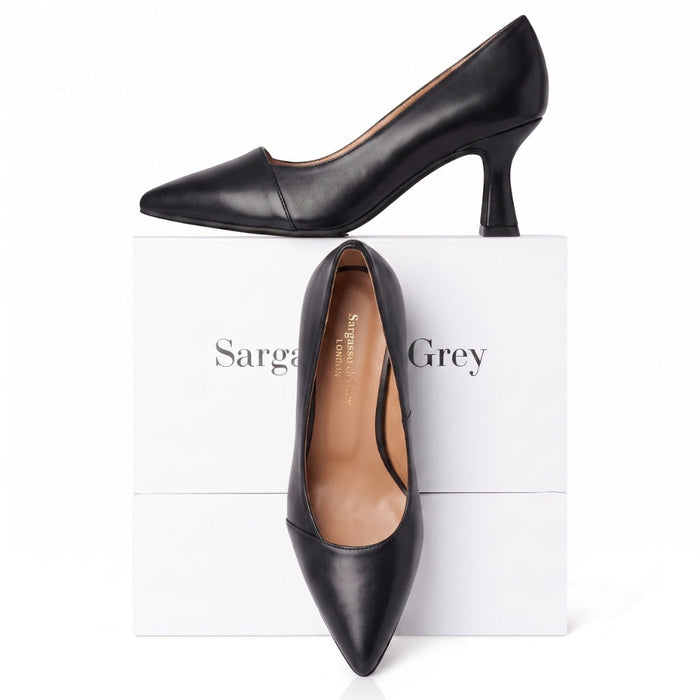 A pair of black pointed-toe kitten heel pumps displayed on top of and in front of a white Sargasso Grey shoebox against a white background.