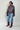 Woman wearing a brown leather jacket, blue jeans, and maroon boots on a plain background