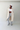 Person wearing a beige trench coat, white top, gray pants, and white sneakers against a plain background