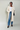 Woman wearing a beige trench coat, white top, blue jeans, and black shoes on a plain background