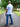 A tall man viewed from behind wearing a plain white short-sleeve t-shirt, blue jeans, and gray sneakers, standing on a gravel path in a park with green trees.