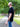 A man wearing a black short-sleeve t-shirt and blue jeans standing on a gravel path in a park with trees. Text on the left reads 'LOW-KEY BRANDING MINIMALIST FRIENDLY' with a white line pointing to the hem wrap logo on the bottom hem.