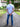 A tall man standing outdoors on a gravel path wearing a white fitted short-sleeve t-shirt and blue jeans, with text overlays highlighting the shirt's features including '100% Mercerised Organic Cotton Planet Friendly' and 'Fitted Sleeves Skinny Arm Fit'.
