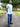 A tall man standing on a park path wearing a white fitted short-sleeve t-shirt and blue jeans, with text callouts describing the shirt's fit and length.