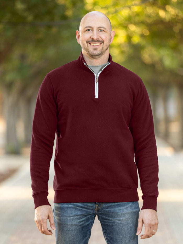 A tall and slim man standing on a path in  a park. The smiling model is wearing a maroon quarter zip sweatshirt. The tall maroon quarter zip sweatshirt features 2.5" longer sleeves, 100% organic french terry cotton, and is soft & comfy. It's  ideal for tall slim men 6'2"+.