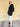 A side-on view of a tall and athletic guy wearing an XL tall black hoodie, standing in front of a beige wall, making hands in pockets and smiling.